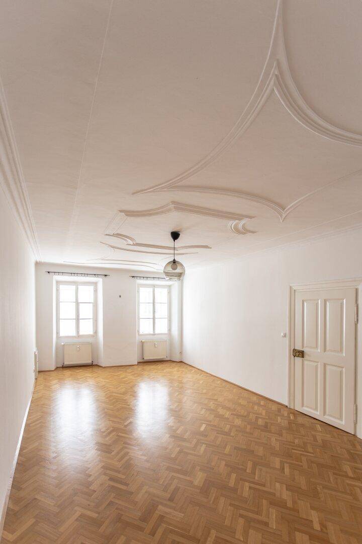 Heller Raum mit Fischgrätparkett, Stuckdecke und zwei großen Fenstern, die viel Licht hereinlassen.