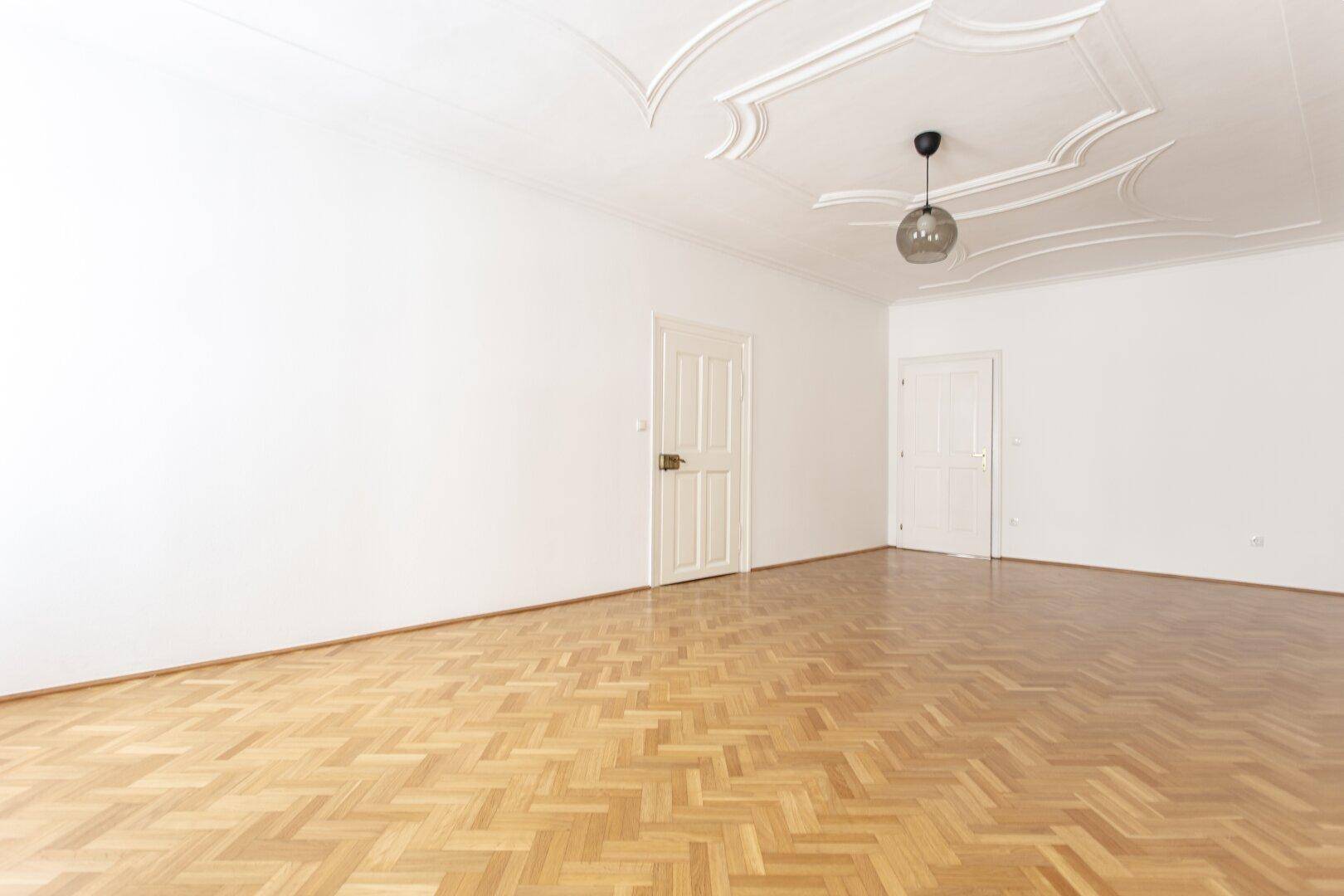 Großes, leeres Zimmer mit Fischgrätparkett und dekorativer Stuckdecke, bereit zur Einrichtung.