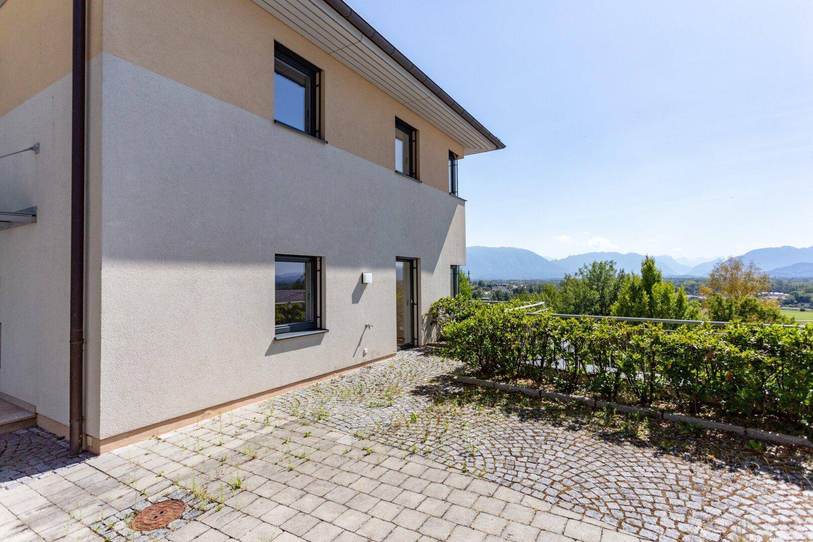 Seitenansicht des Gebäudes mit gepflastertem Bereich und Blick auf die umliegende Landschaft.