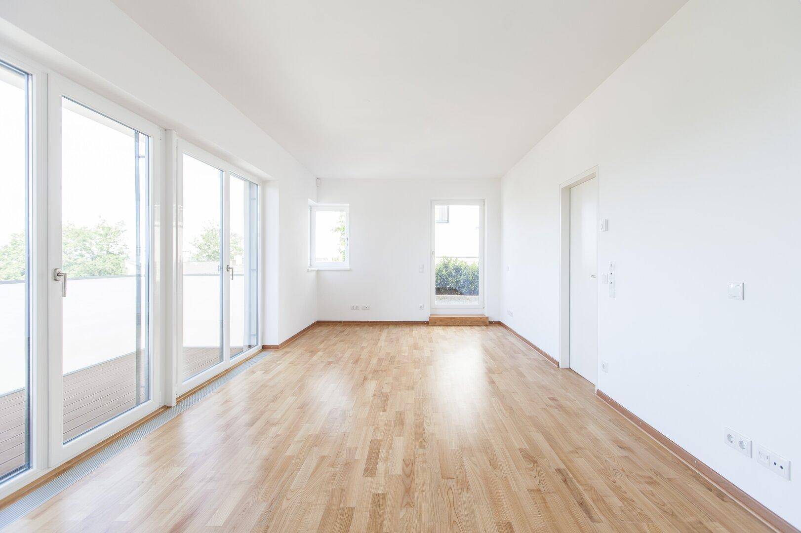 Heller, leerer Wohnraum mit großen Fenstern, Holzboden und Zugang zu einer Terrasse.