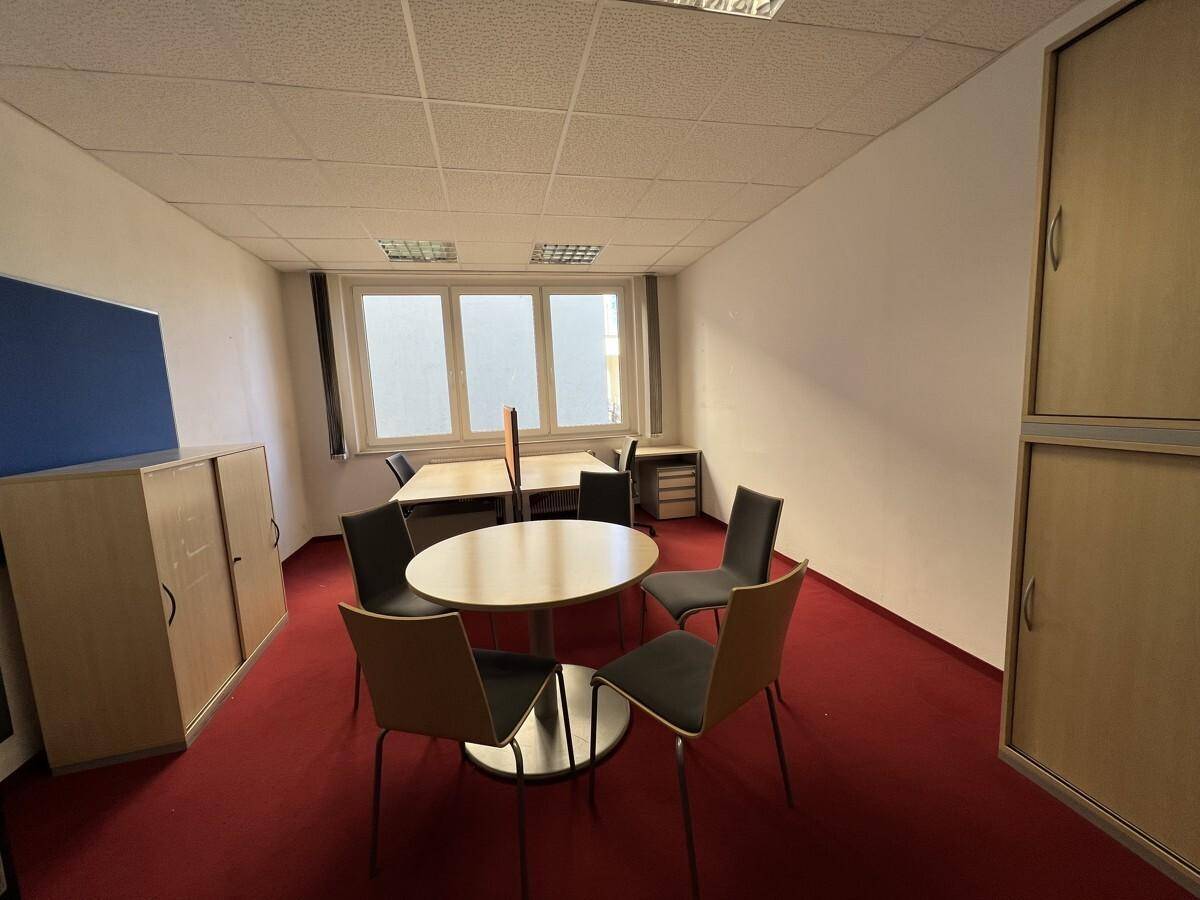Bürofläche mit Besprechungstisch, Schränken und rotem Teppichboden, gut beleuchtet.