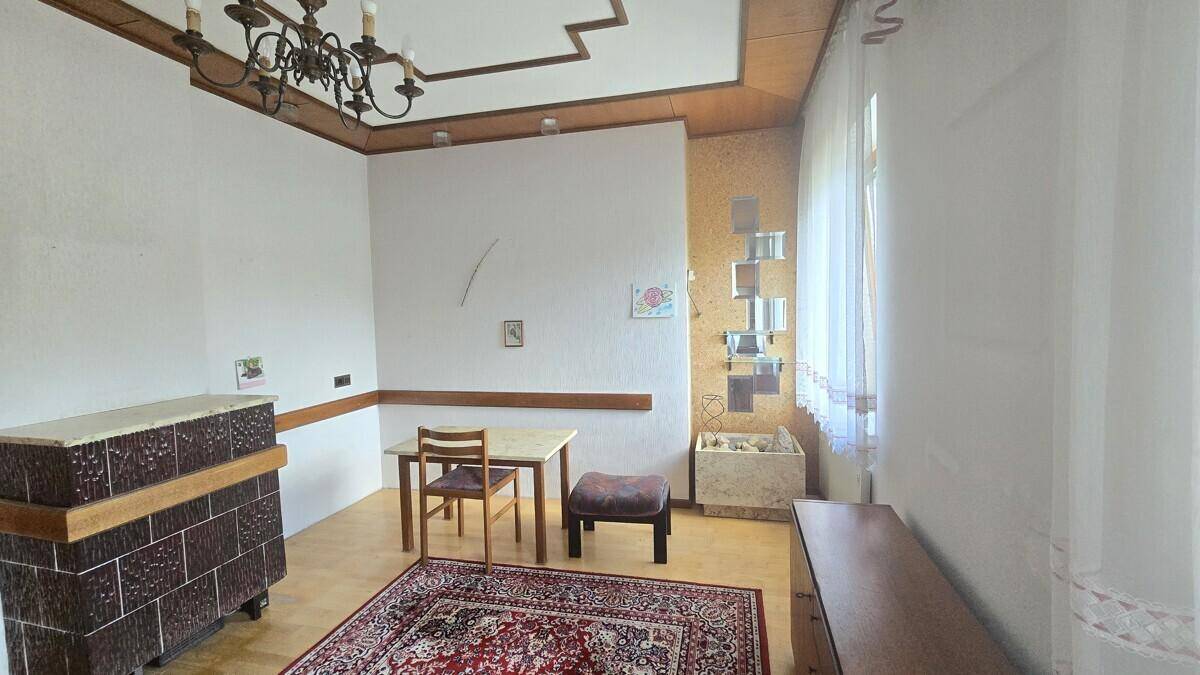Wohnzimmer mit Kachelofen, hellem Holzboden und einem kleinen Esstisch, gemütlich eingerichtet.