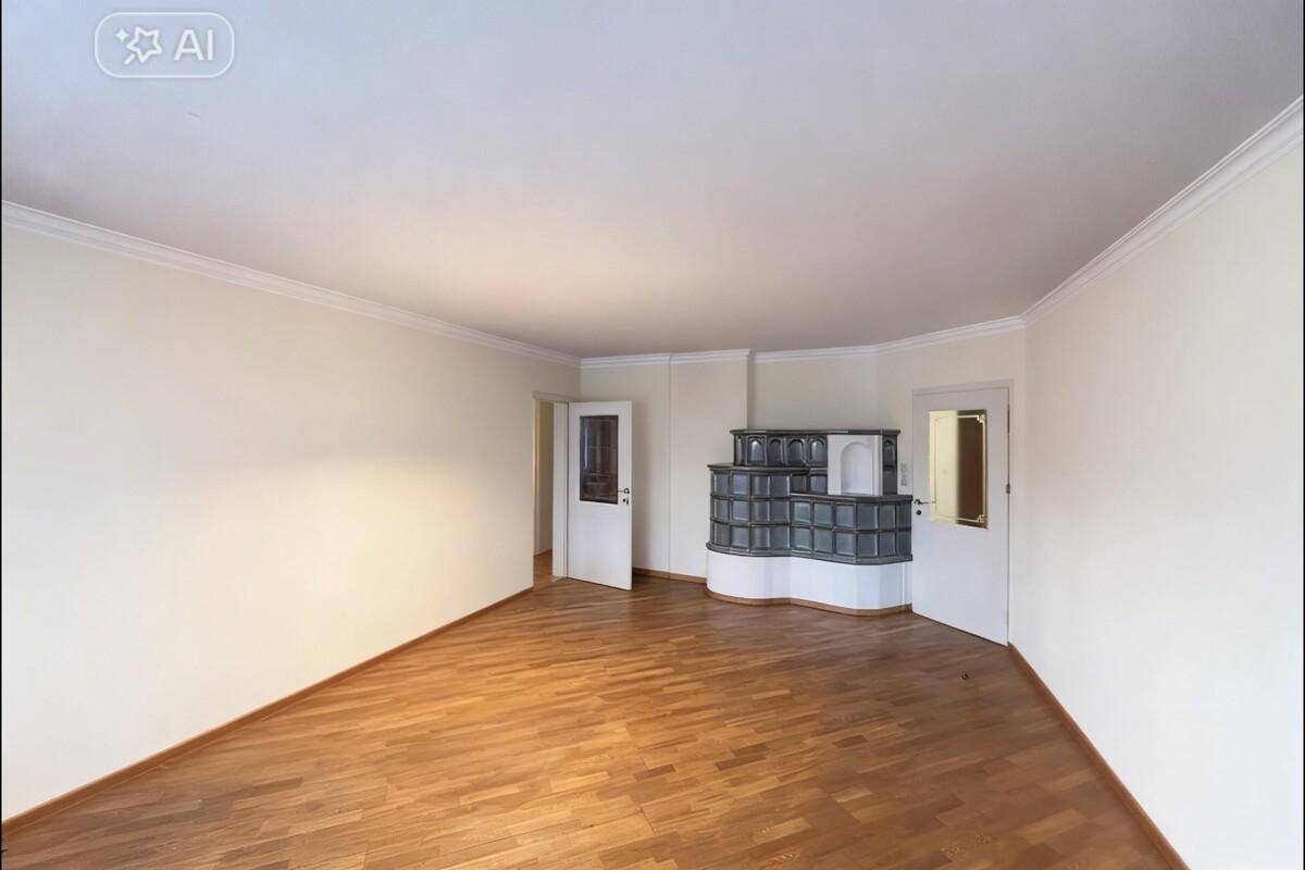 Helles, leeres Wohnzimmer mit Parkettboden und einem traditionellen Kachelofen.