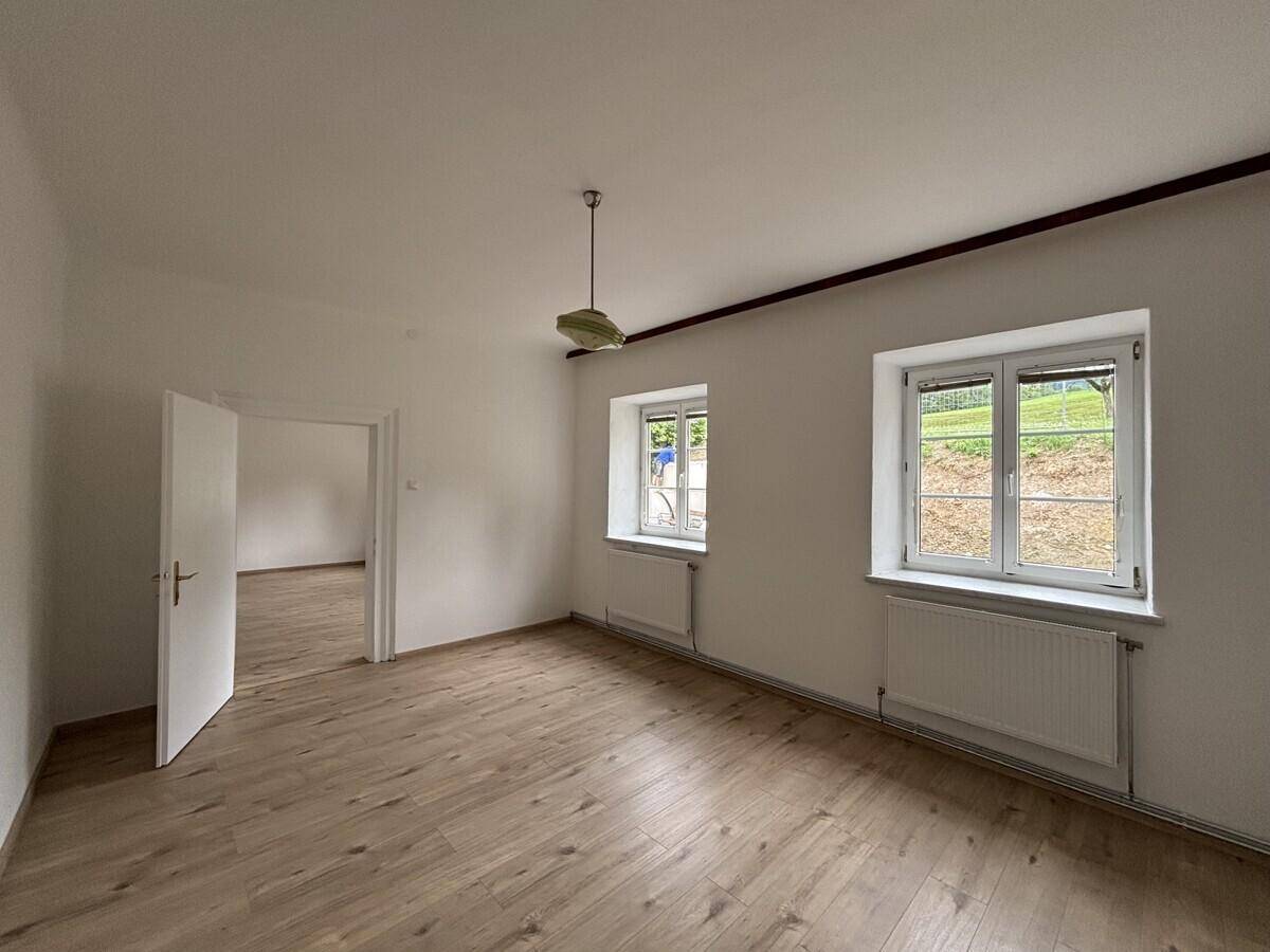Helles Zimmer mit Laminatboden, zwei Fenstern und offener Tür zu einem Nebenraum.