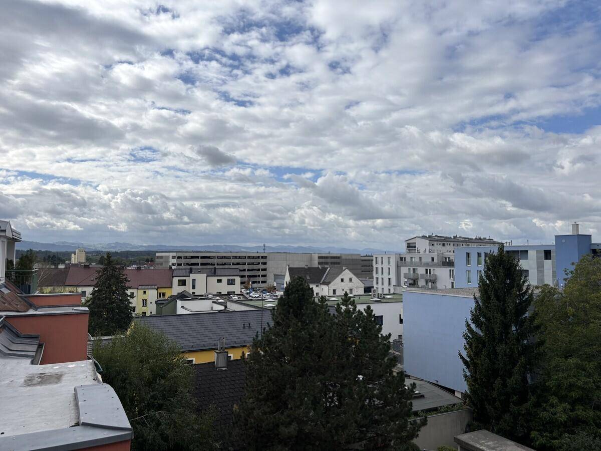 Weitläufiger Blick über die Stadt mit Wohngebäuden, Bäumen und Bergen am Horizont unter bewölktem Himmel.