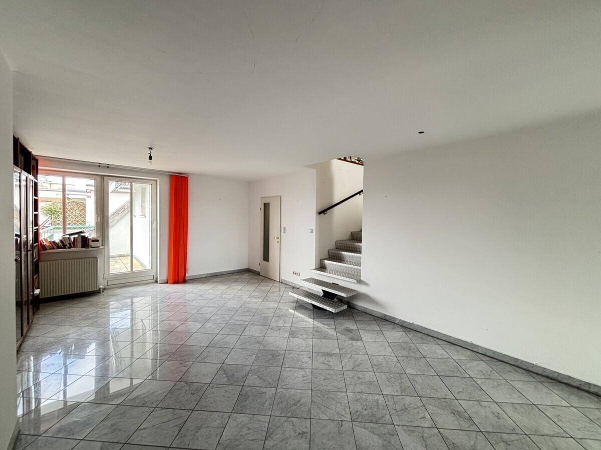 Wohnzimmer mit Zugang zum Balkon, Fliesenboden und offener Treppe.