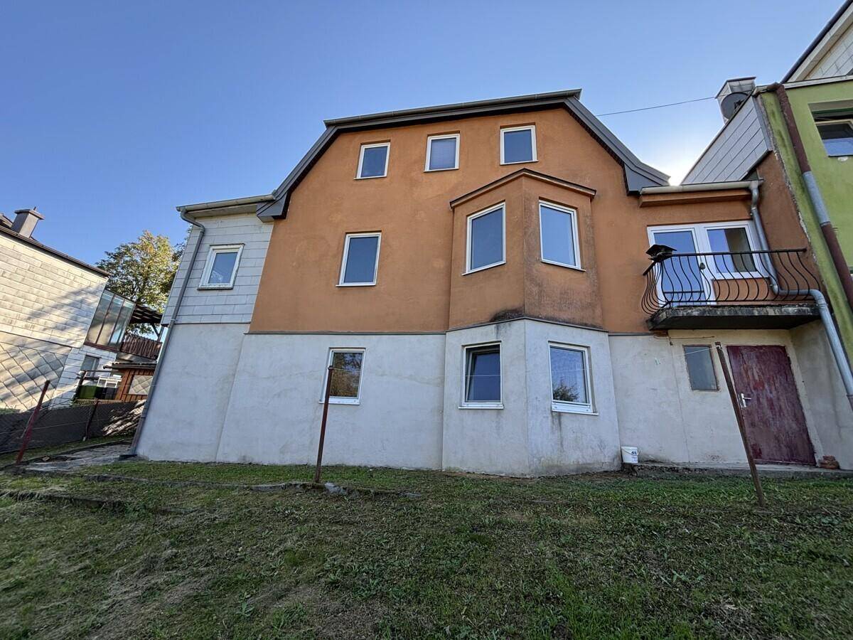 Rückansicht eines Wohnhauses mit mehreren Fenstern und einem kleinen Balkon, umgeben von Grün.
