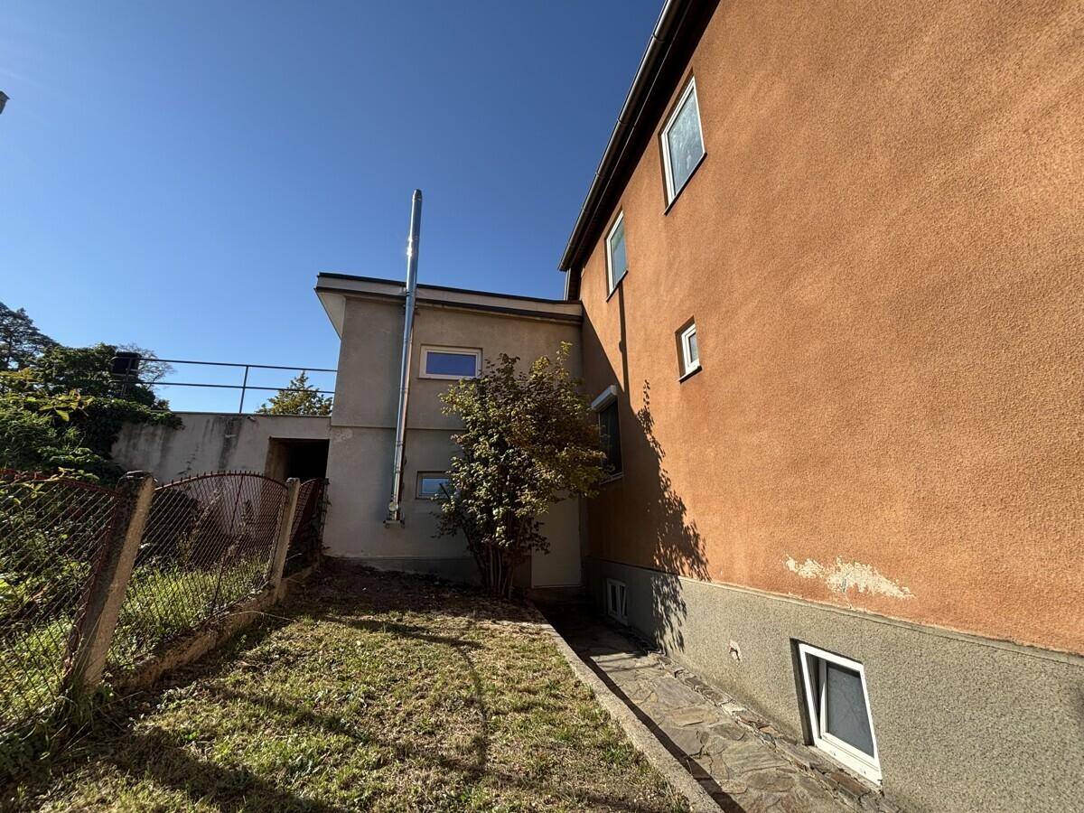 Seitenansicht eines Hauses mit Anbau und einem schmalen Gartenbereich unter blauem Himmel.