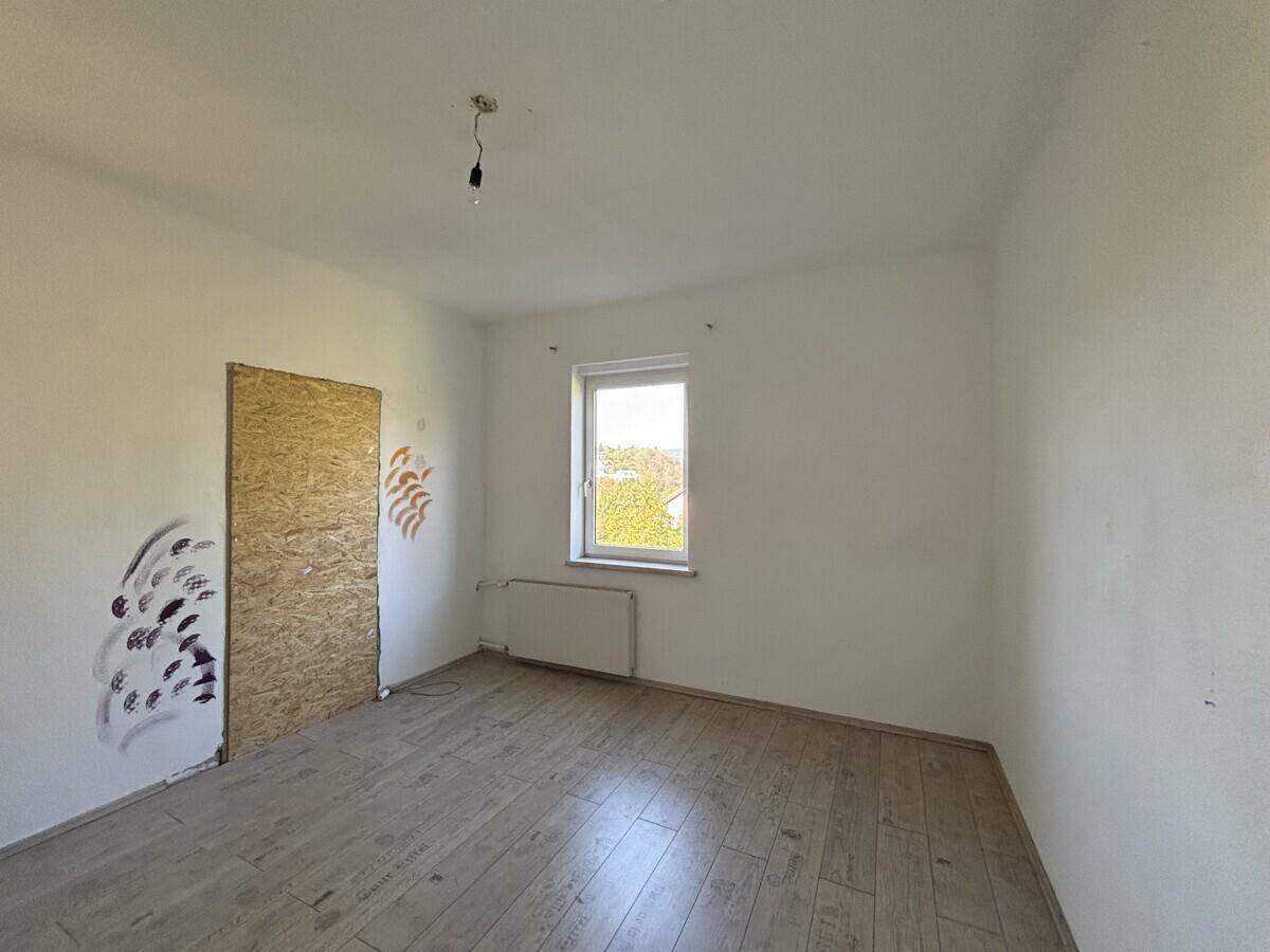 Leeres Zimmer mit Laminatboden, weiß gestrichenen Wänden und einem Fenster mit Ausblick.