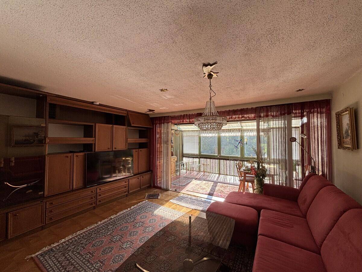 Gemütliches Wohnzimmer mit rotem Sofa, Holzschrankwand und Zugang zu einem hellen Wintergarten.