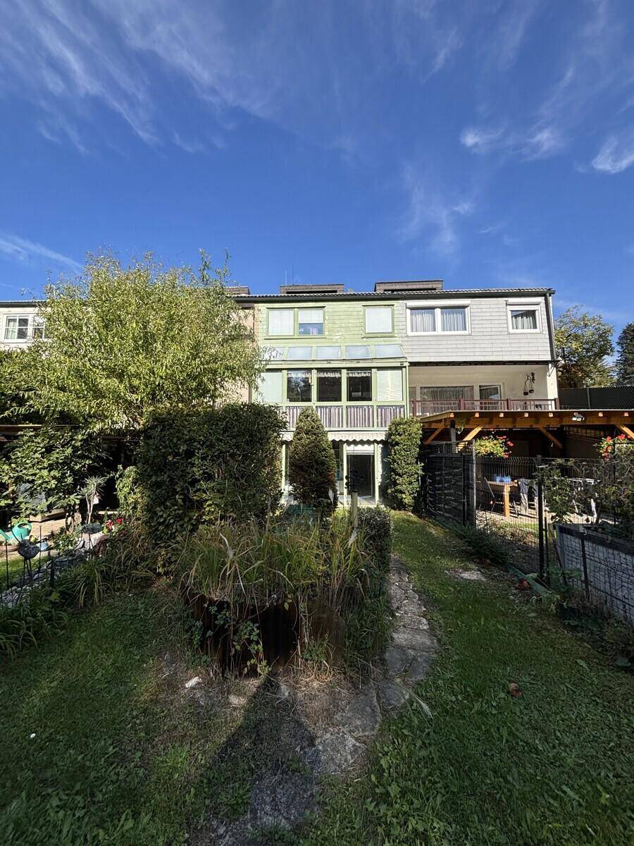 Rückansicht des Reihenhauses vom Garten aus mit Wintergarten und Holzterrasse, umgeben von viel Grün.