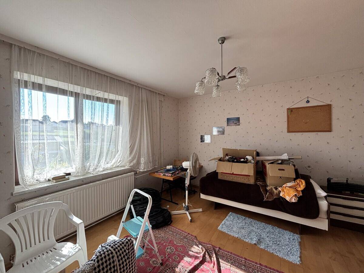 Helles Zimmer mit Holzboden, Fenster und Vorhängen, derzeit als Abstellraum genutzt.