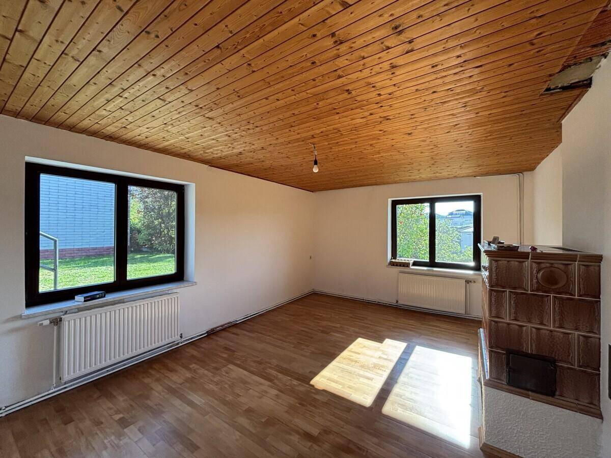 Heller Raum mit Holzboden, Kachelofen und zwei großen Fenstern mit Blick ins Grüne.