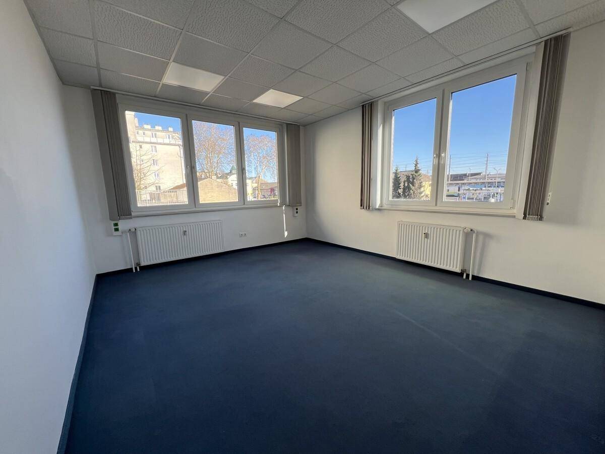 Helles und leeres Büro mit großen Fenstern, blauem Teppichboden und Heizkörpern.