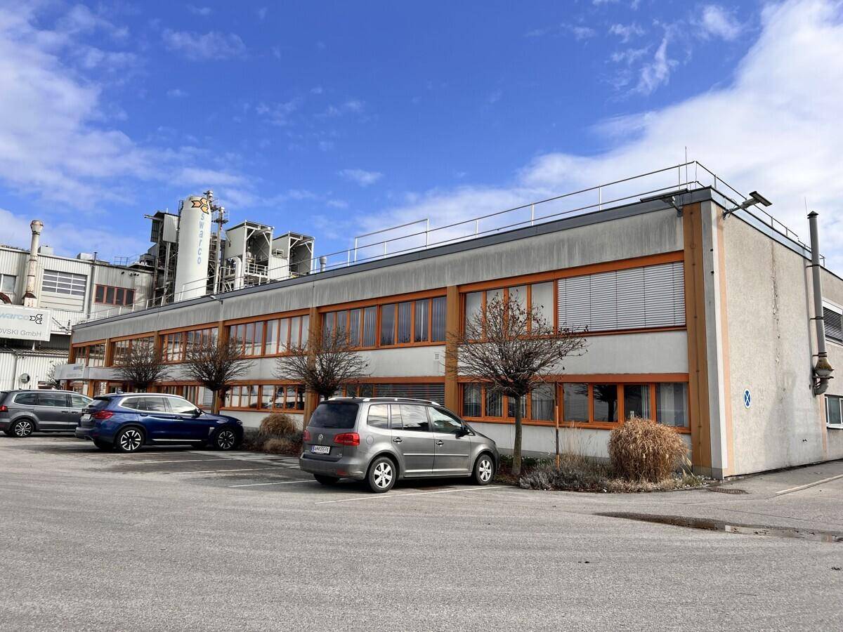 Außenansicht eines Geschäftsgebäudes mit Parkplätzen und industrieller Umgebung im Hintergrund.