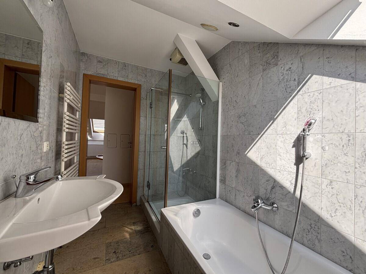 Helles Badezimmer im Dachgeschoss mit Badewanne, Duschkabine, Waschbecken und grauen Fliesen.