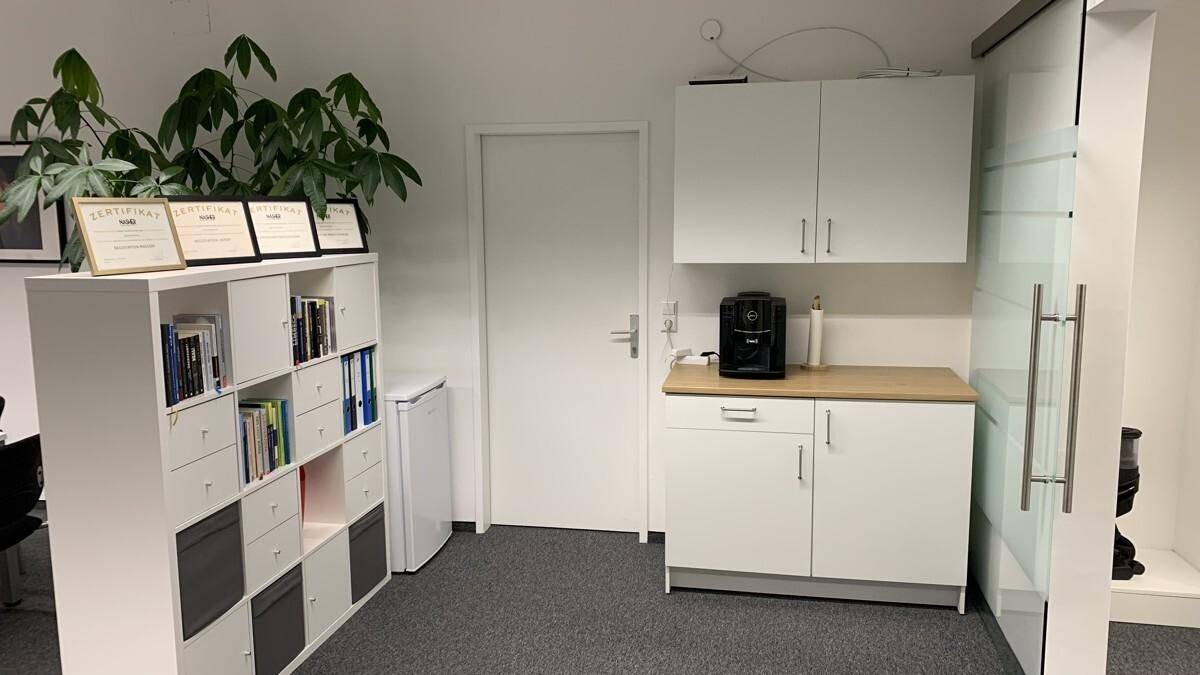 Kleine Küchenzeile in einem Büroraum mit weißen Schränken, Holztheke und Kaffeemaschine.
