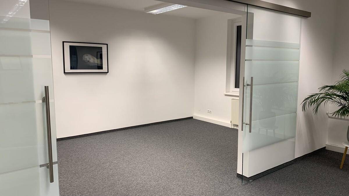 Leerer Büroraum mit Teppichboden, weißen Wänden und einer eleganten Glasschiebetür.