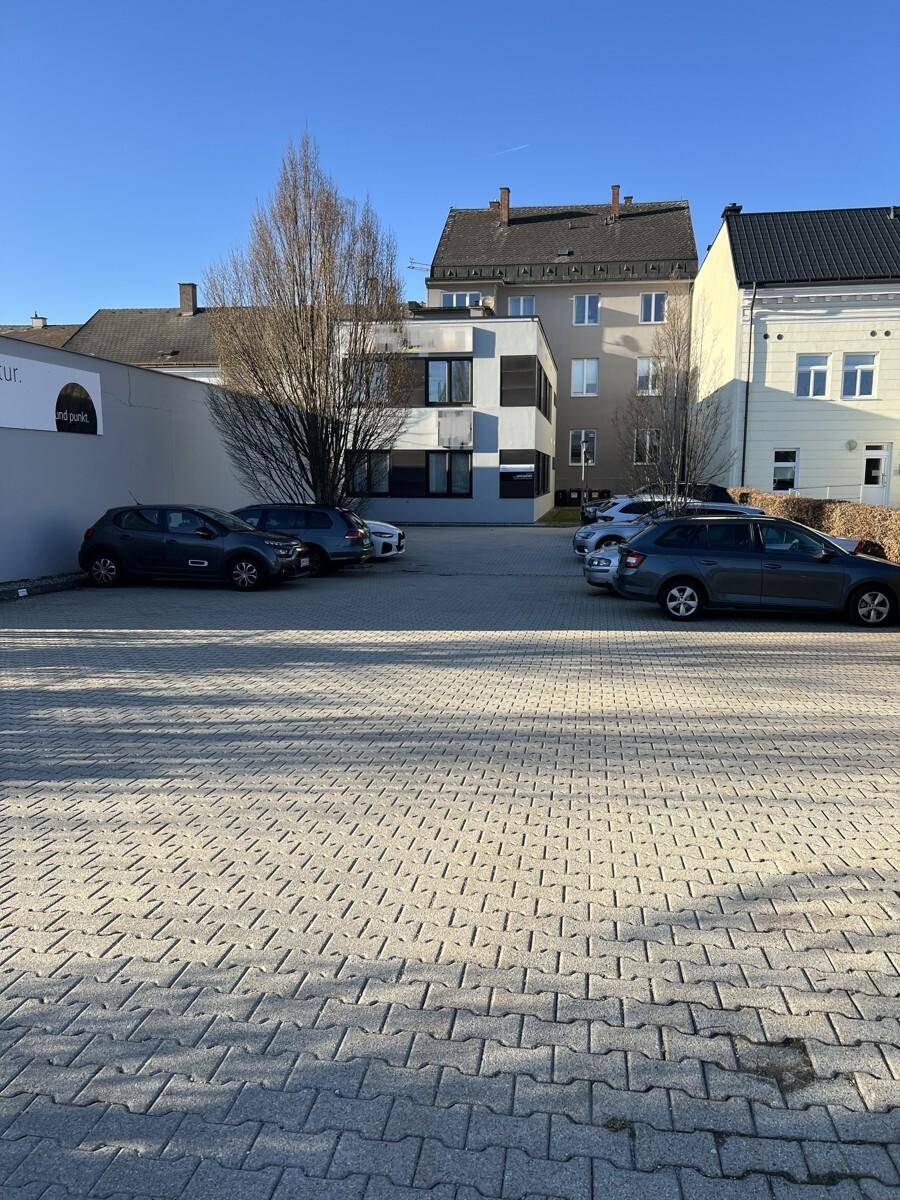 Großer gepflasterter Parkplatz mit mehreren Autos und umliegenden Gebäuden im Hintergrund.