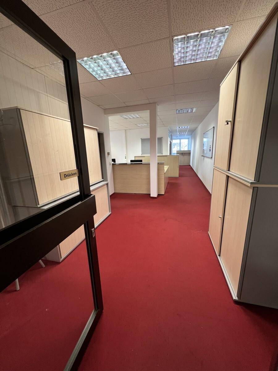 Blick durch eine Glastür in einen langen Büroflur mit rotem Teppichboden und Schränken.