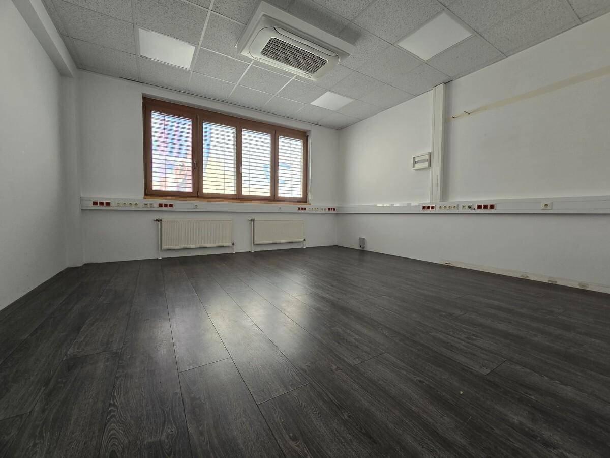 Kleinerer Büroraum mit dunklem Laminatboden, Fenster mit Jalousien und Klimaanlage an der Decke.