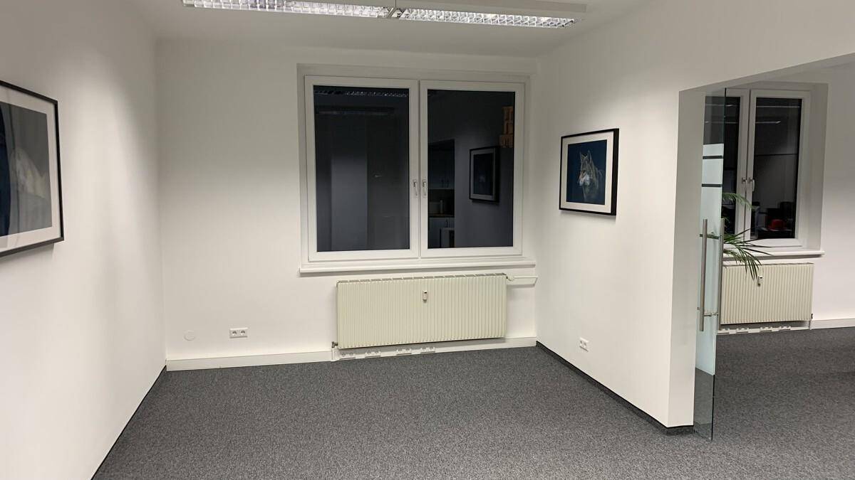 Leerer Büroraum mit großen Fenstern, Heizkörpern und Teppichboden in neutralem Grau.