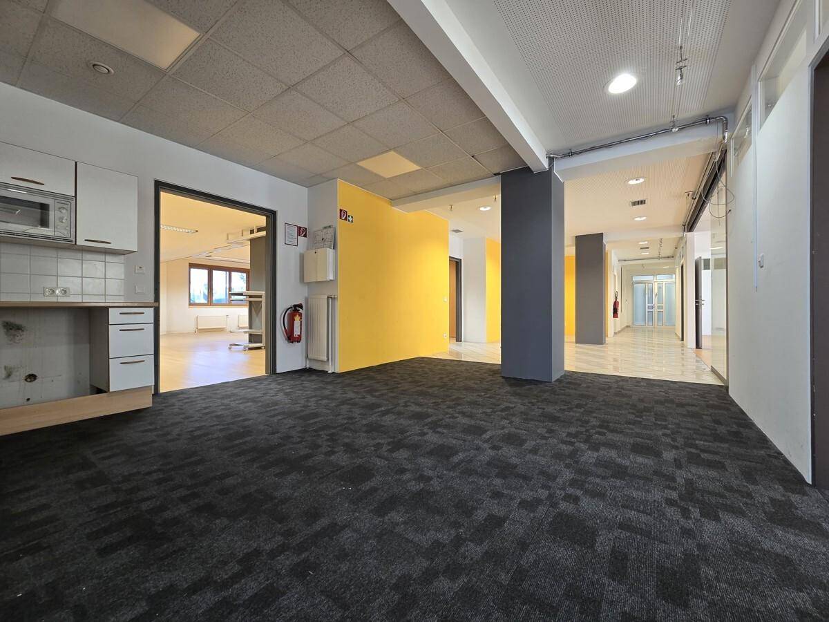 Gewerbefläche mit Teppichboden, kleiner Küchenzeile und Zugang zu weiteren Räumen, mit gelber Wand.