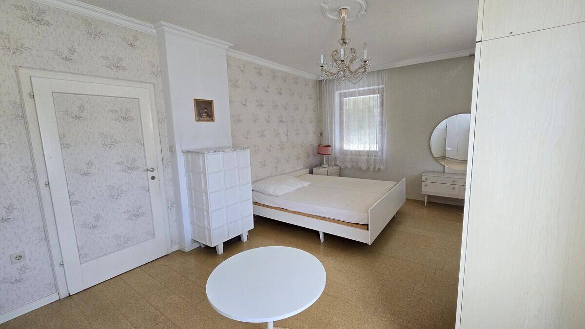 Schlafzimmer mit gemusterter Tapete, hellem Boden, einem Kronleuchter und einem Schminktisch.