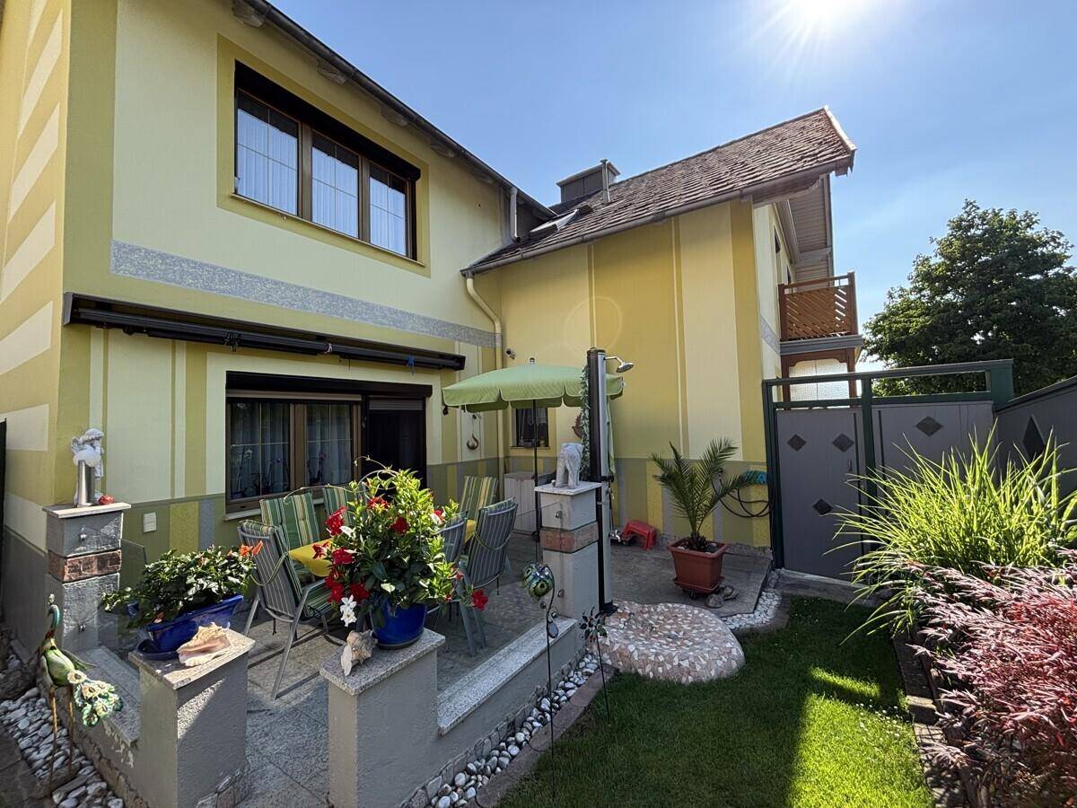 Sonnige Terrasse mit Gartenmöbeln, Sonnenschirm und Blumen vor dem Haus.