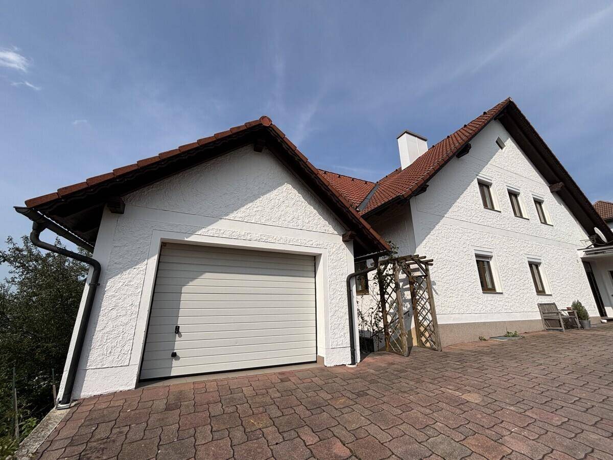 Seitenansicht des Hauses mit integrierter Garage und gepflasterter Auffahrt unter blauem Himmel.