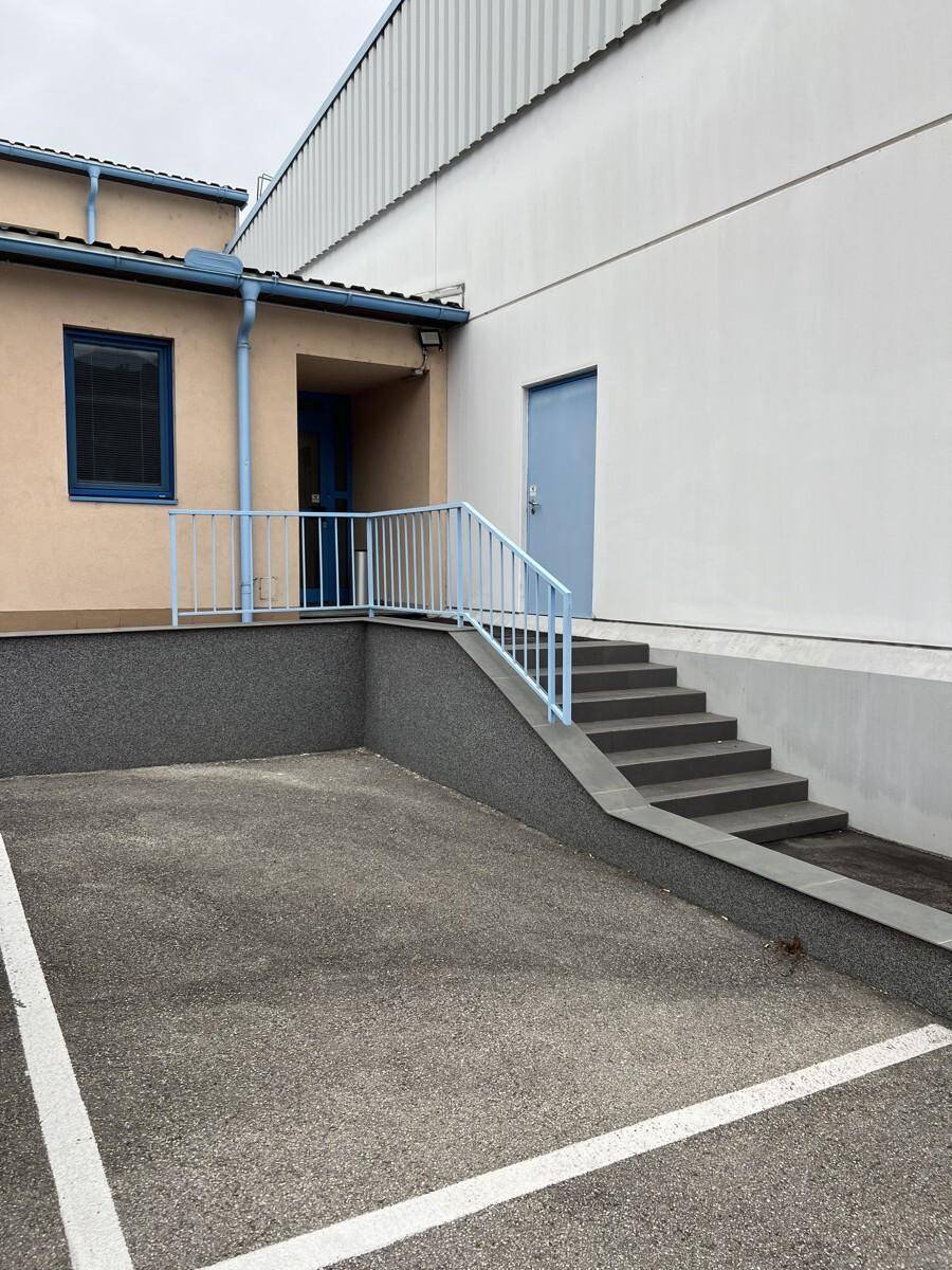 Gebäudeeingang mit Treppe und Geländer, daneben ein Parkplatzbereich.