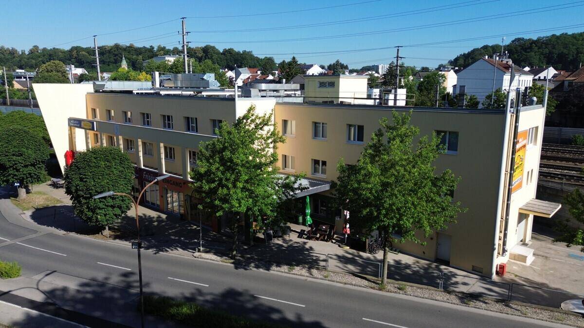 Seitenansicht eines Geschäftsgebäudes mit gelber Fassade, Bäumen und einer Straße im Vordergrund.
