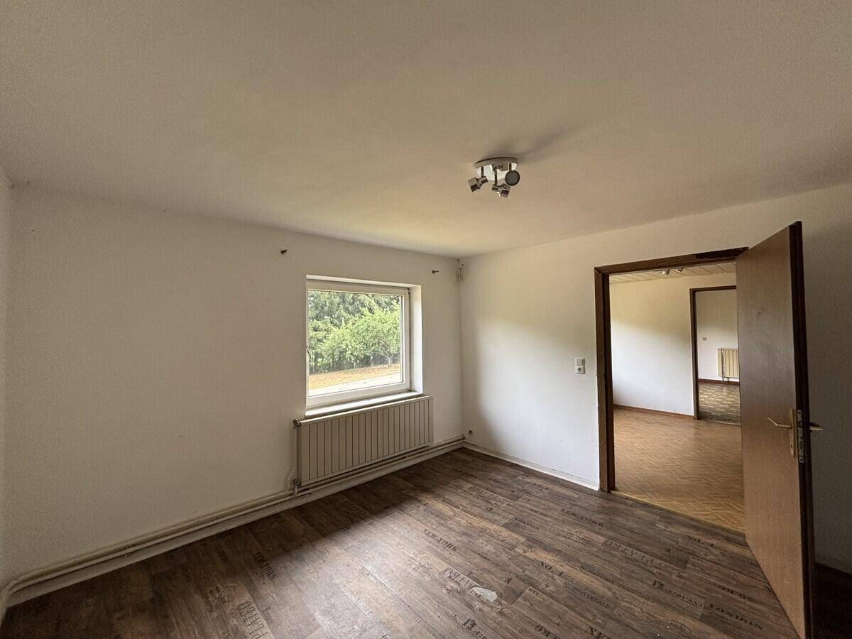 Helles Schlafzimmer mit dunklem Laminatboden, weißen Wänden und Fenster mit Waldblick.