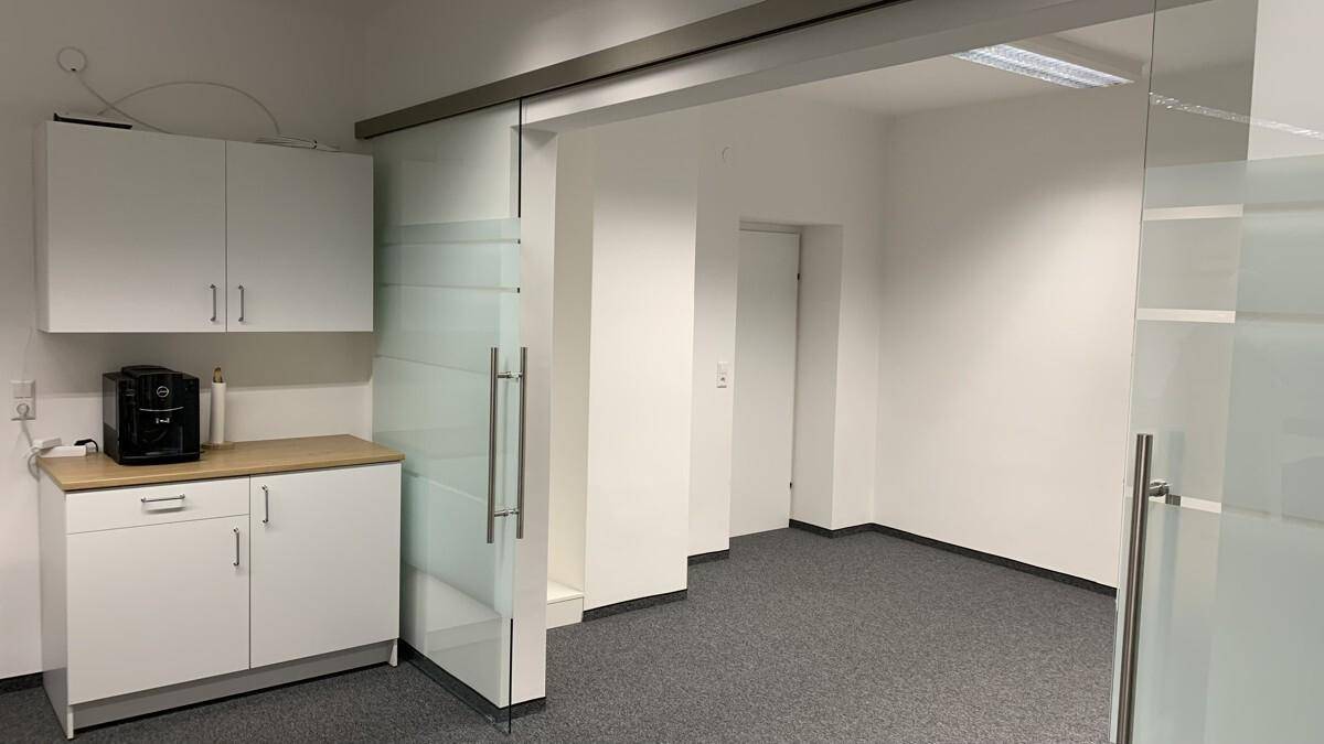 Heller Büroraum mit Teppichboden, weißen Wänden und einer modernen Glasschiebetür.