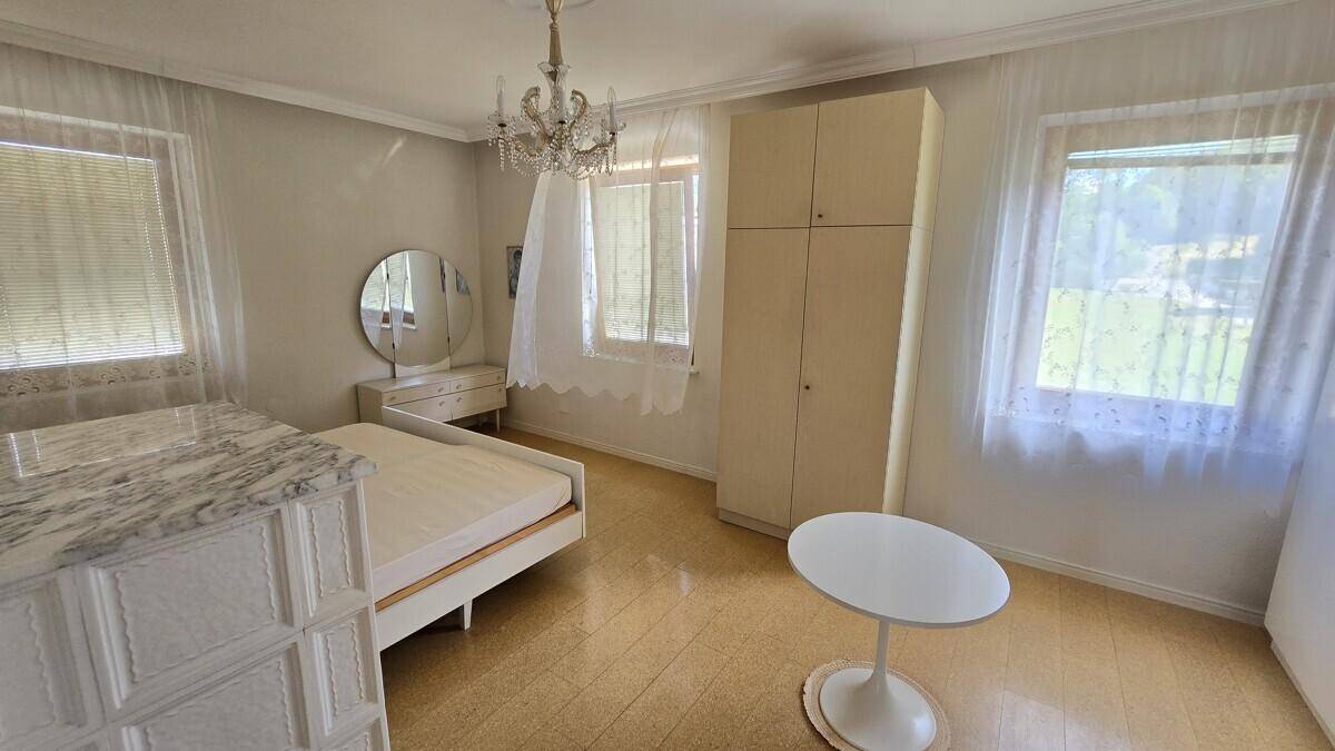 Schlafzimmer mit zwei Fenstern, hellem Boden, einem Kronleuchter und einem Kleiderschrank.