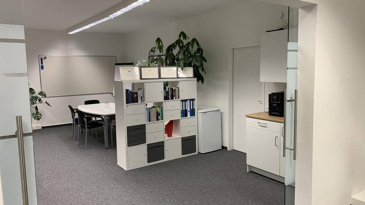 Bürobereich mit Konferenztisch, Regalsystem und einer kleinen Küchenzeile mit Kühlschrank.