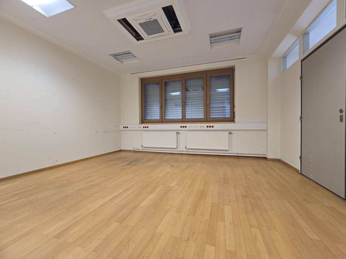 Heller Büroraum mit Holzboden, großem Fenster mit Jalousien und Klimaanlage an der Decke.