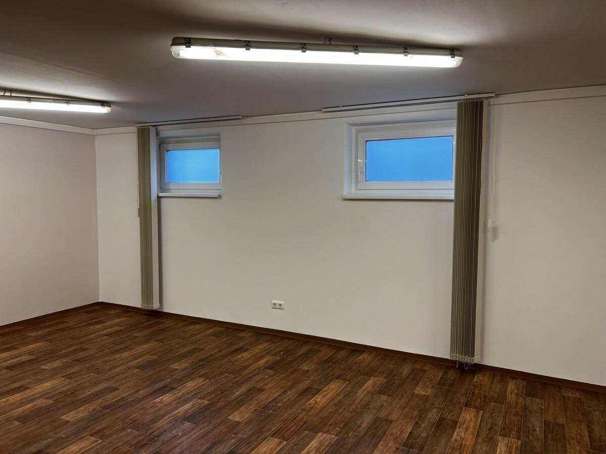 Weiterer Blick in den hellen Raum mit zwei kleinen Fenstern, dunklem Boden und Deckenbeleuchtung.