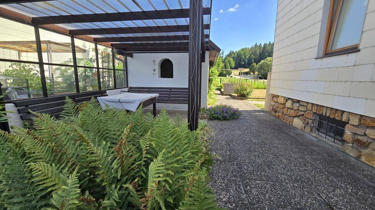 Überdachte Terrasse mit Sitzgelegenheiten, umgeben von Pflanzen und Blick ins Grüne.