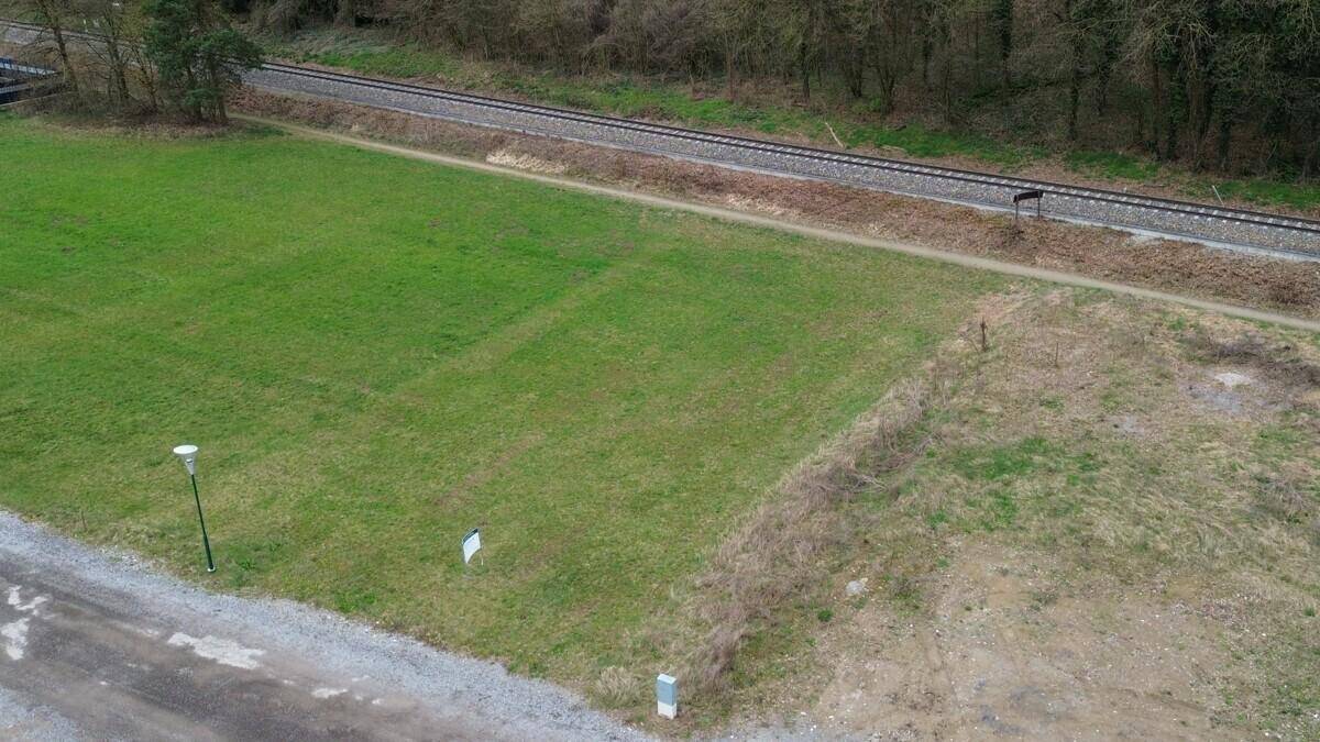 Luftaufnahme eines grünen Grundstücks neben Bahngleisen und einem Waldgebiet mit Schotterweg.