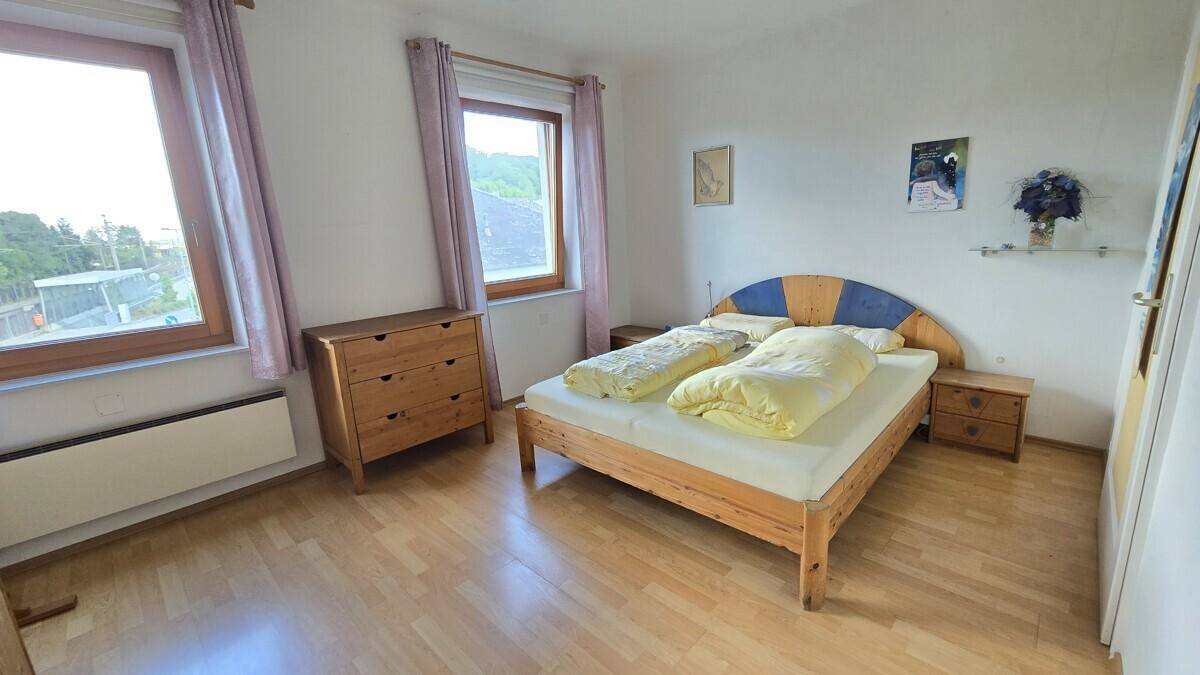 Schlafzimmer mit Holzbett, Nachttischen und zwei Fenstern, die viel Tageslicht hereinlassen.