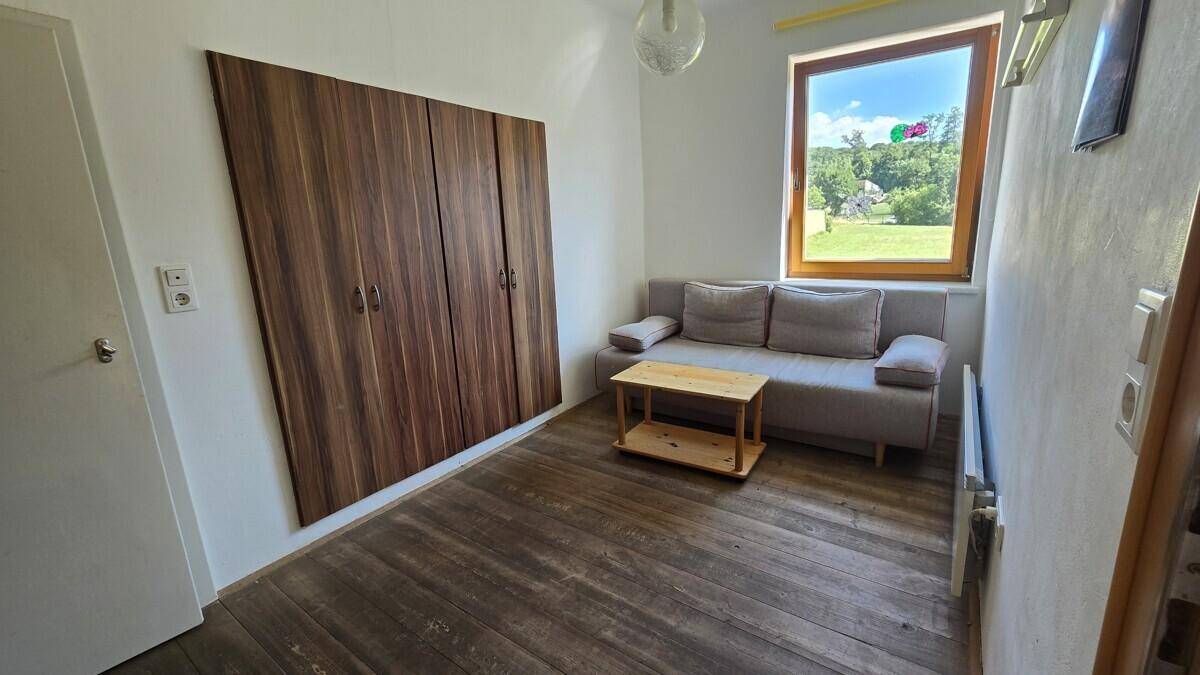 Kleines Zimmer mit Holzboden, einem Sofa, einem großen Holzschrank und Blick ins Grüne.