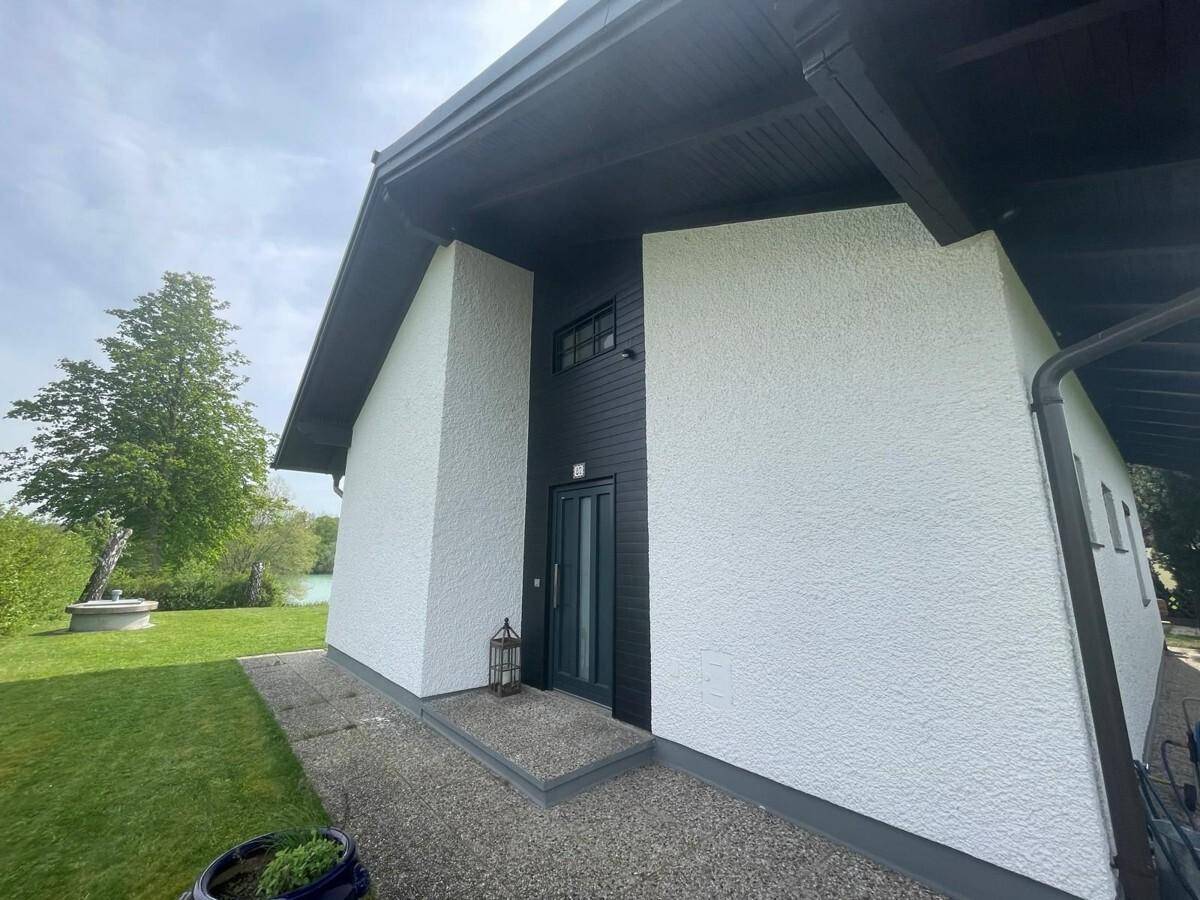 Moderner Hauseingang mit dunkler Holztür und weißer Putzfassade, umgeben von Grün.
