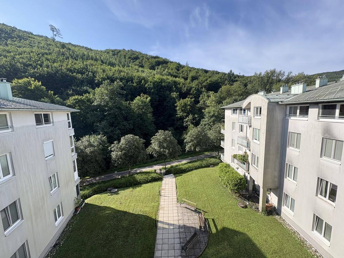 Blick von einem Balkon auf gepflegte Grünflächen zwischen Wohngebäuden und bewaldeten Hügeln.