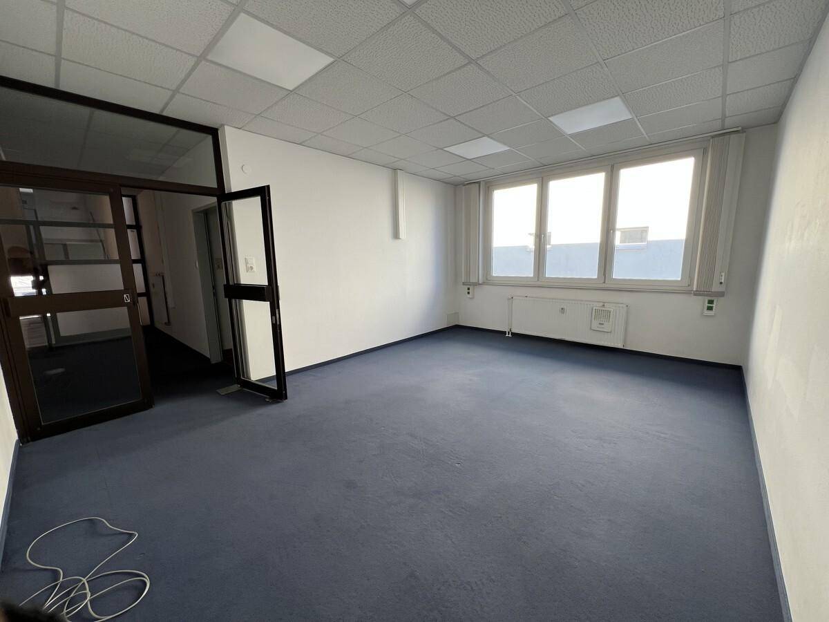 Geräumiges Büro mit Teppichboden, großen Fenstern und Zugang zum Flur.