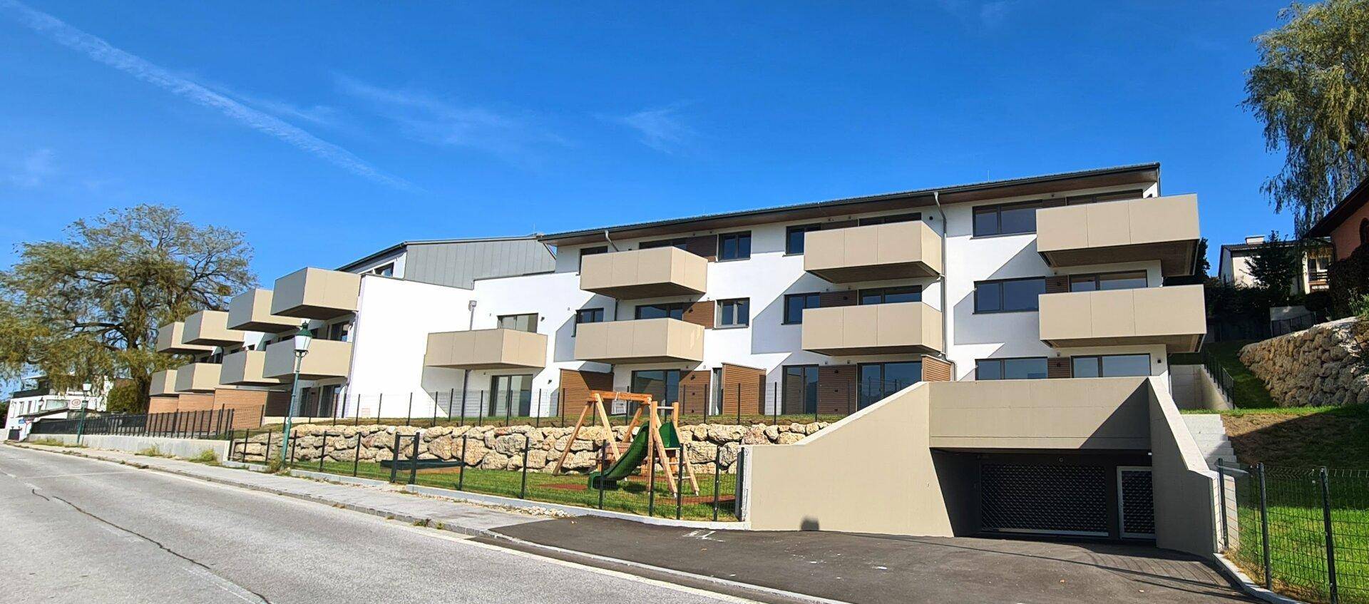 Modernes Mehrfamilienhaus mit weißer Fassade, Balkonen und einem kleinen Spielplatz im Vordergrund.
