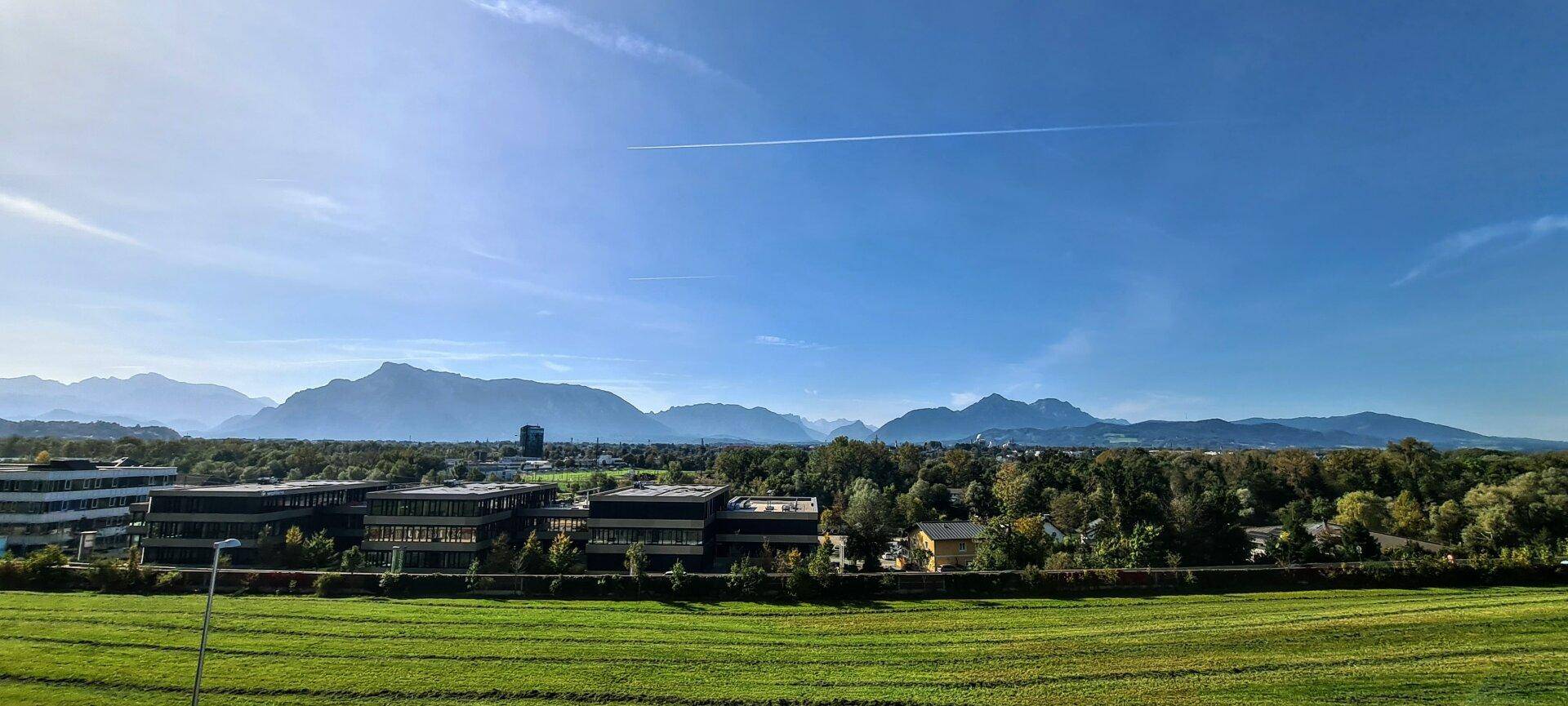 Panoramablick über weite grüne Felder, Gebäude und eine majestätische Bergkette bei strahlendem Sonnenschein.