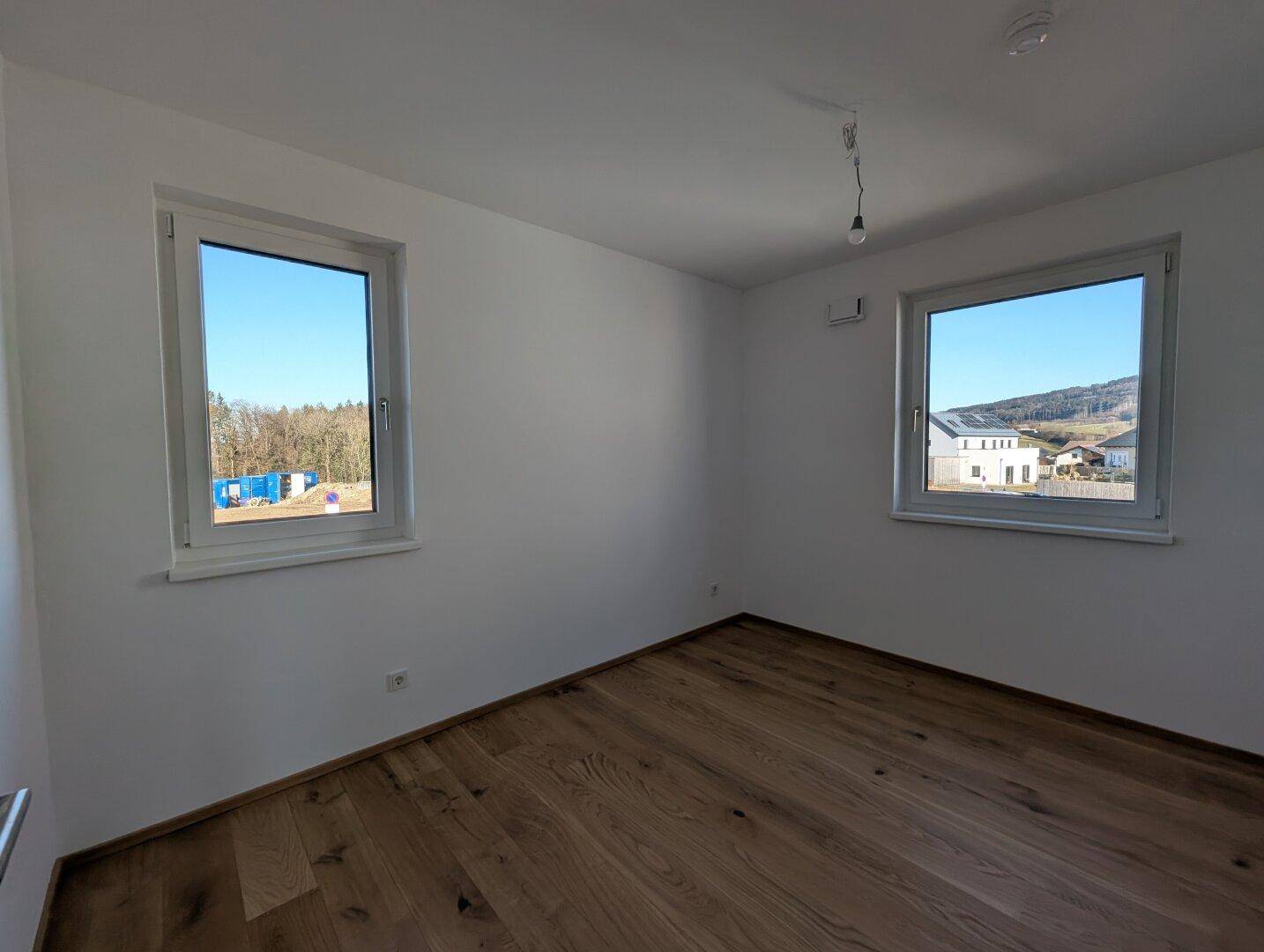 Helles Schlafzimmer mit zwei Fenstern, Eichenparkett und Blick auf die grüne Umgebung.