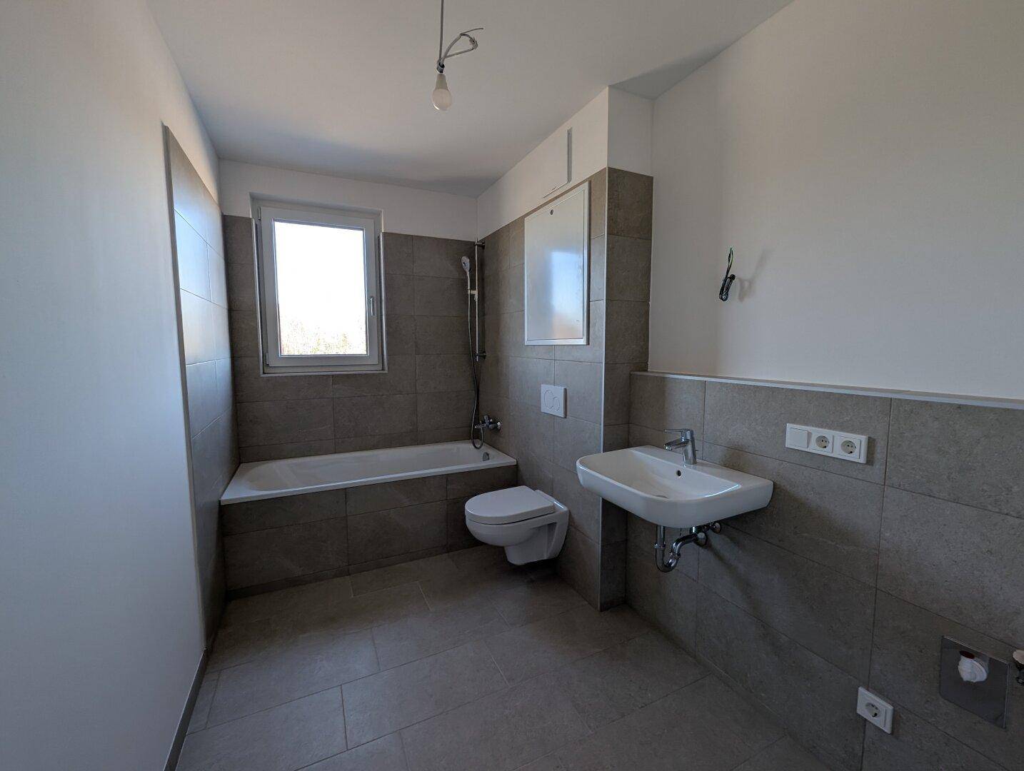 Modernes Badezimmer mit Badewanne, WC, Waschbecken und grauen Fliesen an Wänden und Boden.