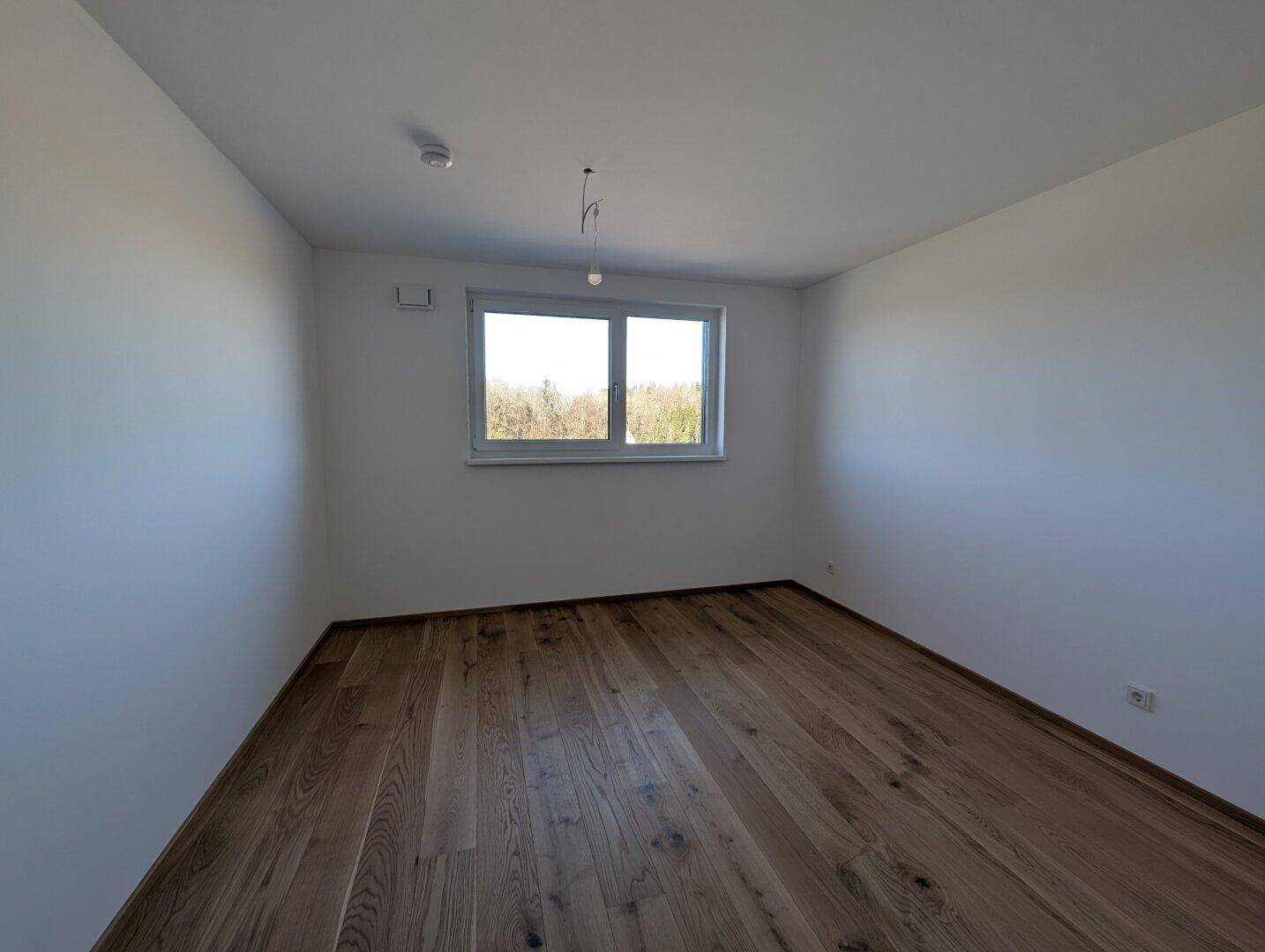 Leeres Schlafzimmer mit Holzboden und großem Fenster, das viel Tageslicht hereinlässt.